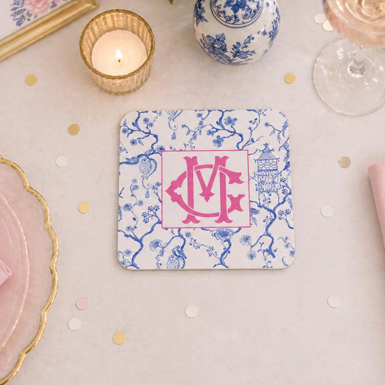 May include: Square white coaster with a blue floral pattern and a pink monogram. The coaster has a pagoda and floral design. A lit candle, a glass of wine, and decorative items are in the background.