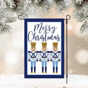 May include: A white garden flag with a blue border and the text "Merry Christmas" in blue script. Four blue and white nutcracker figures stand in a row below the text.