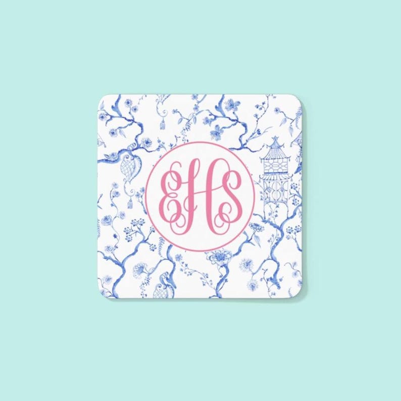 May include: Square coaster with a white background featuring a blue floral pattern and a pink monogram. The monogram is in a pink circle and features the letters EHS.