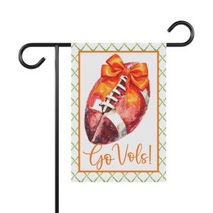 Puede incluir: Una bandera de jard&iacute;n con una ilustraci&oacute;n de acuarela de un bal&oacute;n de f&uacute;tbol americano con un lazo naranja. La bandera tiene las palabras "Go Vols!" en escritura naranja. Est&aacute; bordeada con un patr&oacute;n verde y naranja y montada en un soporte de metal negro.