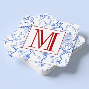 May include: A stack of square coasters with a white background and a blue floral pattern. The top coaster features a red letter "M" inside a red border. The coasters are stacked on a light blue surface.