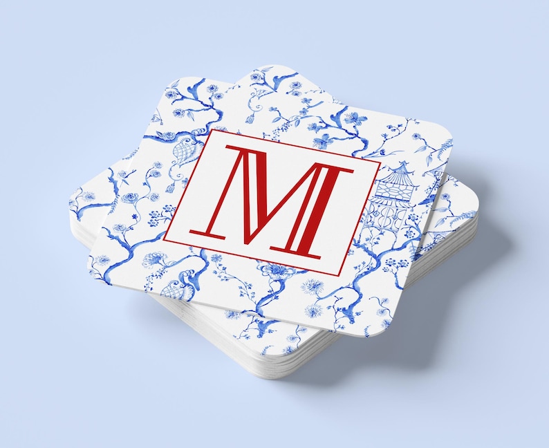 May include: A stack of square coasters with a white background and a blue floral pattern. The top coaster features a red letter "M" inside a red border. The coasters are stacked on a light blue surface.
