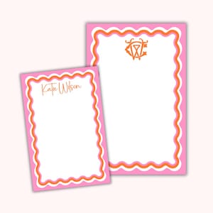 May include: Two pink notepads with orange and white wavy borders. One notepad has the name "Katie Wilson" in orange script. The other notepad has an orange monogram. Both notepads have a white interior.