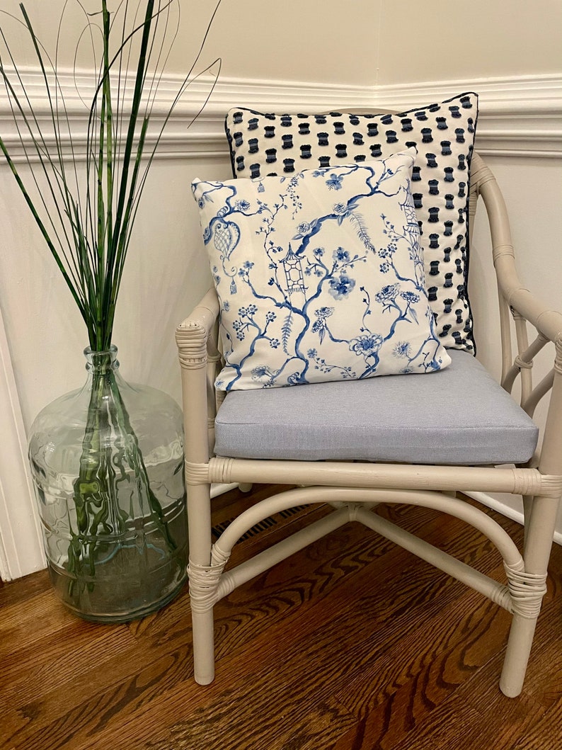 May include: A white wicker chair with a blue and white floral patterned pillow and a black and white patterned pillow. The chair is sitting on a wooden floor.