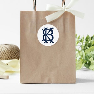 May include: A brown paper gift bag with a white ribbon and a round sticker with the letters "KB" in a navy blue monogram design.