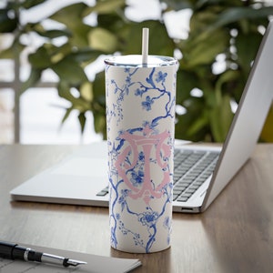 May include: A white stainless steel tumbler with a blue and white floral pattern and a pink monogram. The tumbler has a straw and is sitting on a wooden table.