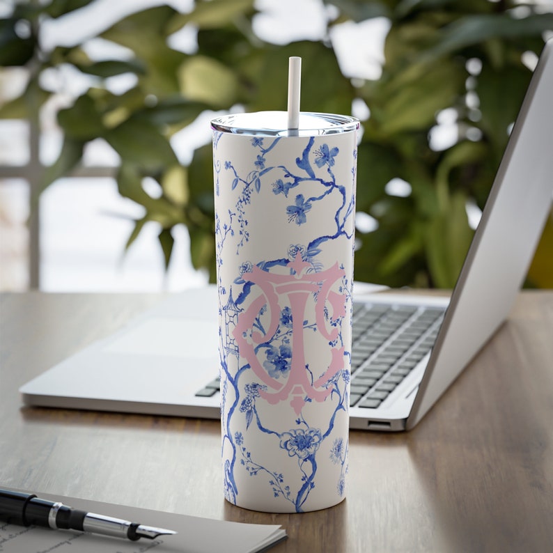 May include: A white stainless steel tumbler with a blue and white floral pattern and a pink monogram. The tumbler has a straw and is sitting on a wooden table.