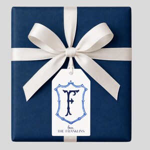 May include: A navy blue gift box wrapped with a white satin ribbon and bow. A white gift tag with a blue crest design and the letter "F" is attached. The tag reads "love, THE FRANKLINS".