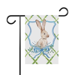 Peut inclure: Un drapeau de jardin blanc avec un motif à carreaux vert et bleu. Un lapin brun avec un nœud bleu se trouve au centre du drapeau. Le lapin est entouré de feuilles vertes et de fleurs bleues.