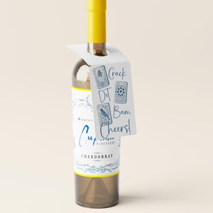 May include: A clear wine bottle with a yellow cap and a white tag. The tag has the words "Crack Dot Bam Cheers!" and blue illustrations. The bottle label says "Cupcake Vineyards Chardonnay".