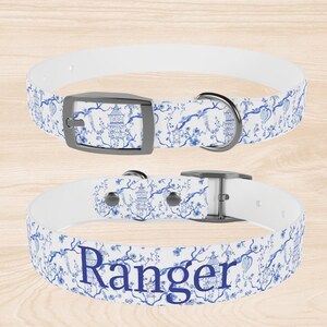 Op de afbeelding: Witte hondenriem met een blauw en wit bloemenpatroon en de naam "Ranger" in blauw bedrukt. De riem heeft een zilveren gesp en een zilveren D-ring.