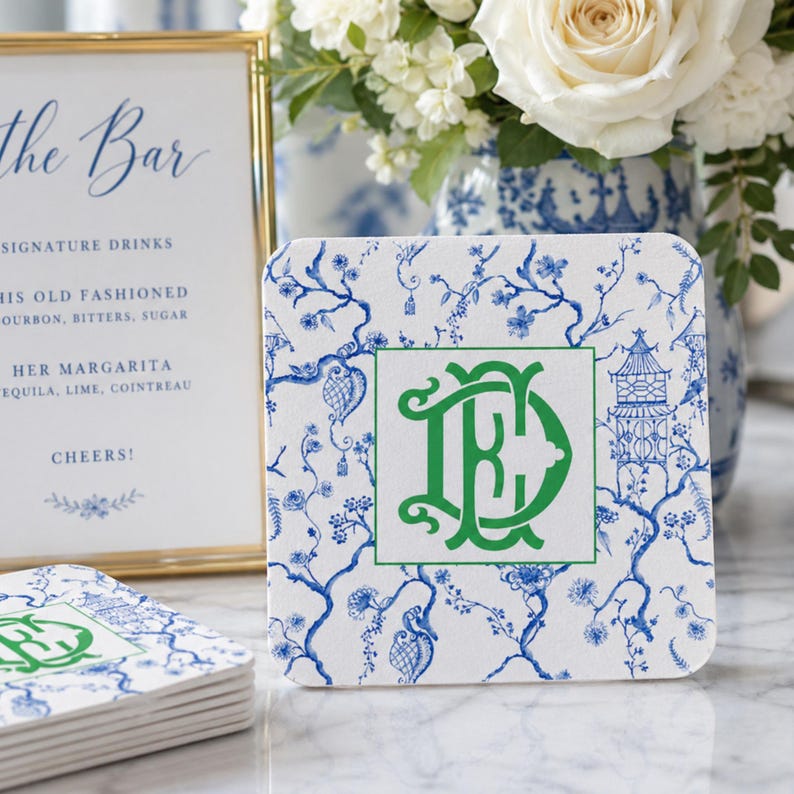 May include: Square white coasters with a blue floral pattern and a green monogram. The coasters are stacked near a sign that says "The Bar" and a vase of white flowers. The coasters are approximately 4 inches square.
