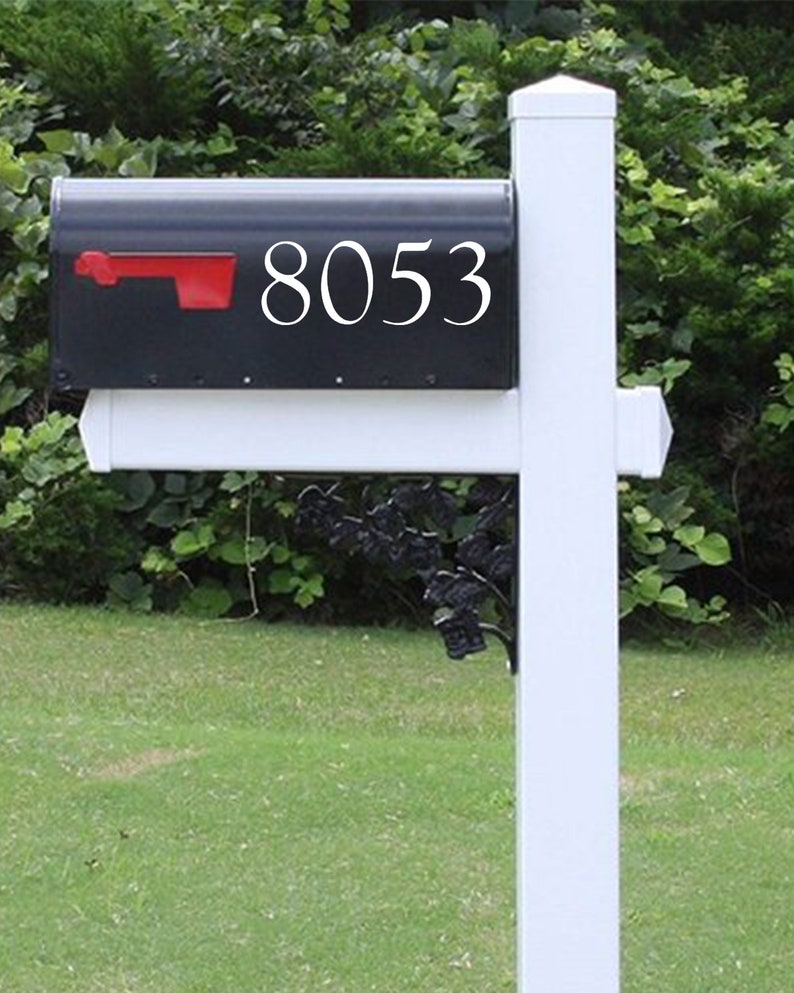 May include: A black mailbox with the number 8053 in white lettering. The mailbox has a red flag and is mounted on a white post. The background features green grass and foliage.