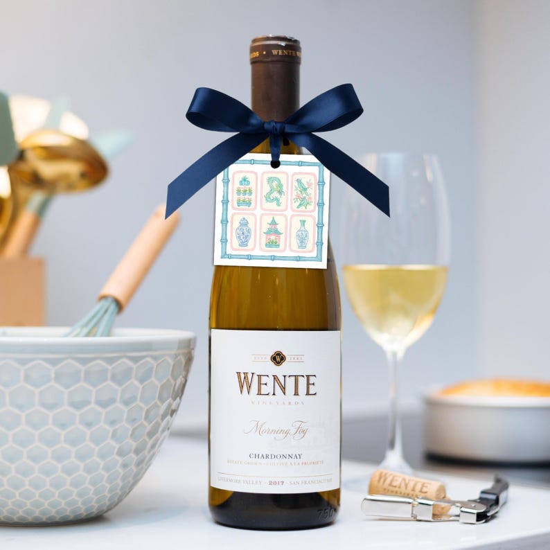 May include: A bottle of Wente Chardonnay wine with a navy blue ribbon and a decorative tag. The tag features a bamboo border and illustrations of vases and other Asian-inspired designs. A wine glass with wine and a corkscrew are also visible.