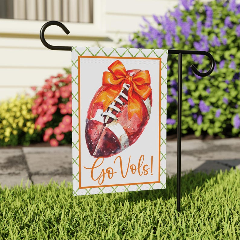 Puede incluir: Una bandera de jard&iacute;n decorativa con un bal&oacute;n de f&uacute;tbol de estilo acuarela y un lazo naranja. La bandera tiene un borde naranja y un patr&oacute;n de trama cruzada verde. Las palabras "Go Vols!" est&aacute;n escritas en naranja.