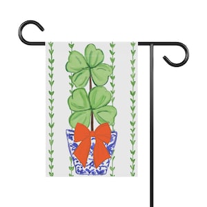 Peut inclure: Un drapeau de jardin avec un fond blanc et une plante de trèfle à quatre feuilles verte dans un pot bleu et blanc avec un nœud rouge. La plante est entourée de vignes vertes.