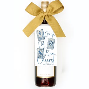 May include: A bottle of red wine with a gold-colored bow and a white tag. The tag features blue and white illustrations and the words "Crack Dot Bam Cheers!" with a signature. The bottle has a gold cap.
