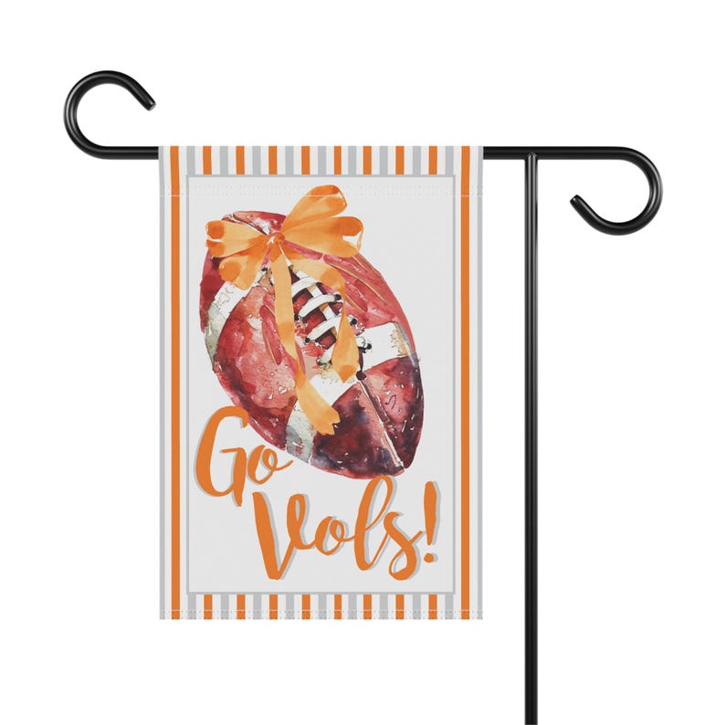 Puede incluir: Una bandera de jard&iacute;n con un bal&oacute;n de f&uacute;tbol en acuarela y un lazo naranja. La bandera tiene las palabras "Go Vols!" en escritura naranja. La bandera est&aacute; bordeada con rayas naranjas y grises y se muestra en un soporte de metal negro.