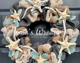 May include: A handcrafted wreath with a coastal theme. It features burlap in shades of gray, tan, and cream, adorned with starfish, rope, and faux greenery. The wreath is displayed on a black door.