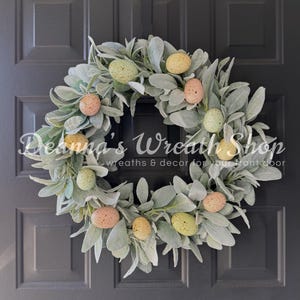 May include: A green lambs ear wreath decorated with pastel colored speckled Easter eggs. The wreath is hanging on a dark gray door.