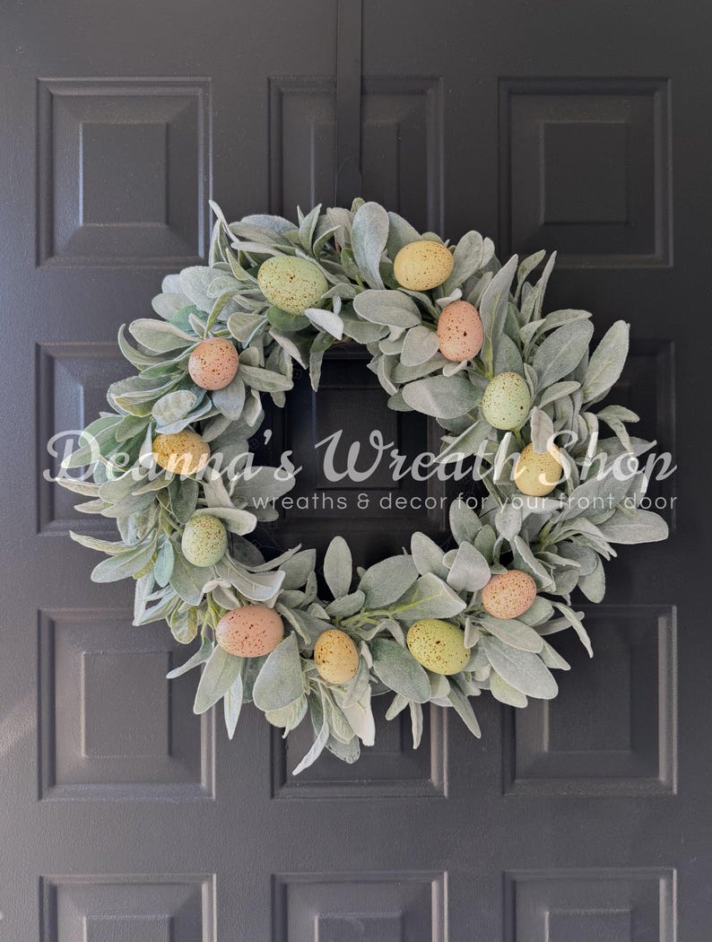 May include: A green lambs ear wreath decorated with pastel colored speckled Easter eggs. The wreath is hanging on a dark gray door.