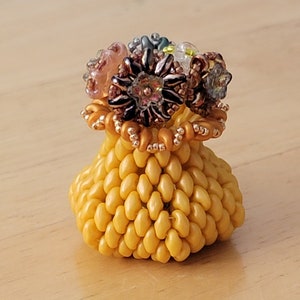 May include: A small, yellow, beaded vase with a beaded rim and a cluster of colorful beaded flowers on top.