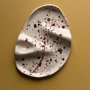 Könnte beinhalten: Eine weiße Keramik-Servierplatte mit organischer Form und drei Abschnitten. Die Platte ist mit dunkelroter Spritzfarbe verziert. Der Hintergrund ist einfarbig senfgelb.