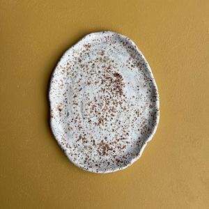 Puede incluir: Plato de cerámica blanca con forma irregular, salpicado de manchas marrones. El plato tiene un aspecto artesanal. Está sobre un fondo amarillo mostaza. El plato mide aproximadamente 25 cm de largo.