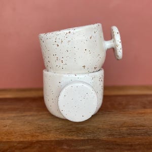 Op de afbeelding: Twee gestapelde, gespikkelde witte keramische botervlootjes. De bovenste schaal heeft een handvat en de onderste schaal heeft een deksel. De schalen staan op een houten oppervlak met een roze achtergrond.