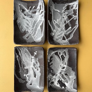 Puede incluir: Juego de cuatro platos rectangulares de cerámica negra con diseños abstractos blancos. Los platos son ligeramente curvos y tienen un aspecto rústico y hecho a mano.