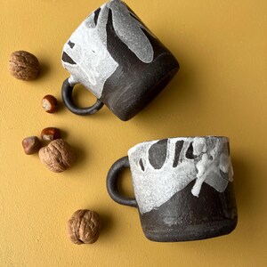 Peut inclure: Deux tasses en céramique noire avec des motifs abstraits blancs. Les tasses ont une forme arrondie et une anse.