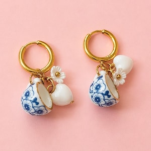 Op de afbeelding: Een paar gouden oorringen met bedels. Elke oorring heeft een miniatuur blauw-wit kopje, een wit hart en een kleine witte bloem. De achtergrond is zachtroze.
