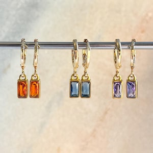 May include: Gold-coloured hoop earrings with rectangular gemstones in orange, blue, and purple. The earrings are suspended from a silver bar. The background is a soft, blurred gradient. The earrings are a jewellery item.