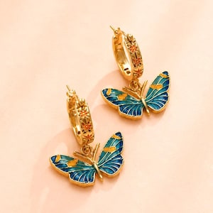 May include: A pair of gold-colored hoop earrings with butterfly pendants. The butterflies have blue and yellow enamel wings. The hoops are decorated with small orange floral designs. The earrings are displayed against a soft pink background.