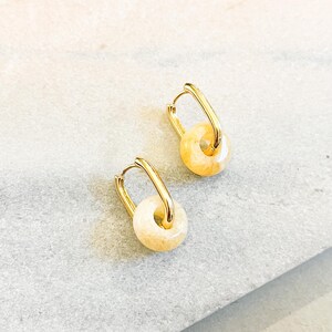 May include: A pair of gold-colored hoop earrings with a small, round, light-yellow stone pendant. The earrings are displayed on a light gray surface.