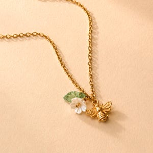 May include: A gold-toned metal chain necklace featuring three charms: a textured gold bee, a small white flower with five petals, and a single green leaf. The delicate chain links are uniform in size and connect to the charms at the bottom centre.