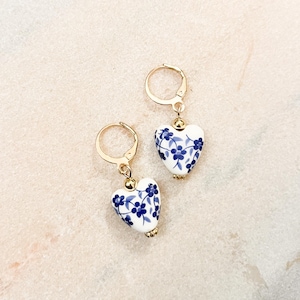 Può includere: Un paio di orecchini a cerchio color oro con ciondoli a forma di cuore. Ogni ciondolo è bianco con un motivo floreale blu. Gli orecchini presentano una piccola perlina color oro sulla parte superiore del cuore dove si collega al cerchio.
