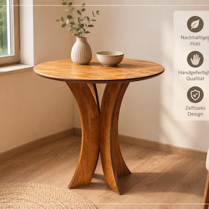 Könnte beinhalten: Ein runder Holztisch mit glatter Oberfläche und einem Fuß aus drei geschwungenen, sich kreuzenden Beinen. Auf dem Tisch stehen eine geriffelte weiße Vase mit einer Grünpflanze und eine kleine, schlichte Schale. Der Text rechts lautet: Nachhaltiges Holz, Handgefertigte Qualität, Zeitloses Design.