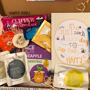 May include: A gift box filled with various items, including Clipper hot chocolate, English Tea Shop tea, a Yankee Candle, face mask, and a card that says "It's just such a good day to bee happy". The box is lined with pink tissue paper.