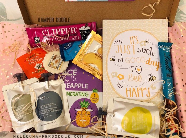 May include: A gift box filled with various items, including Clipper hot chocolate, English Tea Shop tea, a Yankee Candle, face mask, and a card that says "It's just such a good day to bee happy". The box is lined with pink tissue paper.