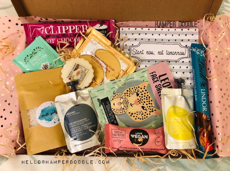 May include: A gift box filled with various self-care items. Includes Clipper hot chocolate, English Tea Shop tea, Yankee Candle, face masks, bath salts, a Lindor chocolate bar, and a Love Vegan bar. The box is lined with pink tissue paper and decorative straw.