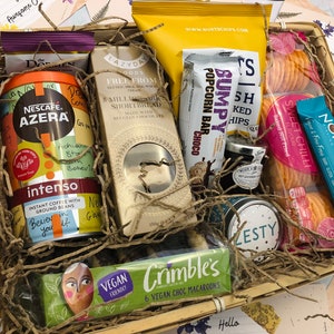 May include: A wicker basket filled with a variety of snacks and treats, including coffee, shortbread, popcorn, crackers, and chocolate. The basket is lined with brown paper shredding and includes a variety of colorful packaging.