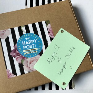 May include: A brown cardboard box with a sticker that says "Yay Happy Post!" and a floral design. A light green note is attached with the words "Enjoy!!" and "Hamper Doddle".