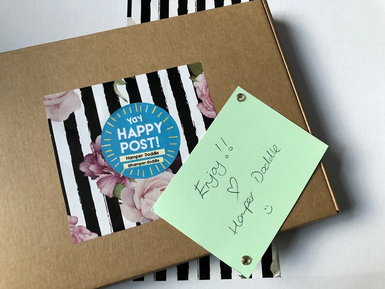 May include: A brown cardboard box with a sticker that says "Yay Happy Post!" and a floral design. A light green note is attached with the words "Enjoy!!" and "Hamper Doddle".