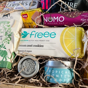 May include: A gift basket filled with various items, including a box of lemon zest cookies, a box of tea, a chocolate bar, a candle, and a small tin of tea. The basket is filled with shredded paper.