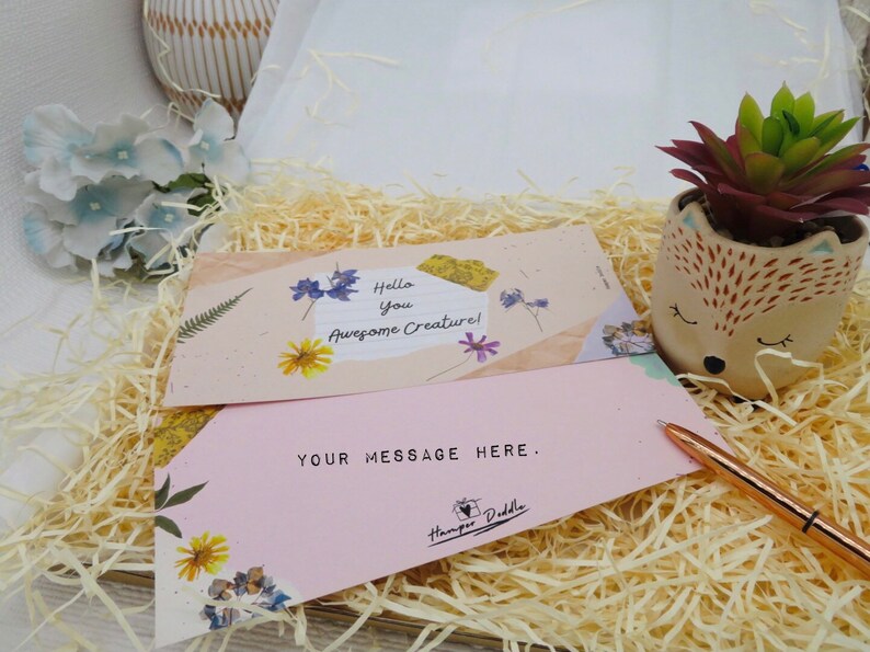 May include: A pink and beige card with the text "Hello you Awesome Creature!" and "YOUR MESSAGE HERE."  The card is decorated with dried flowers and is sitting on a bed of shredded paper. A small succulent plant in a ceramic fox planter is in the background.