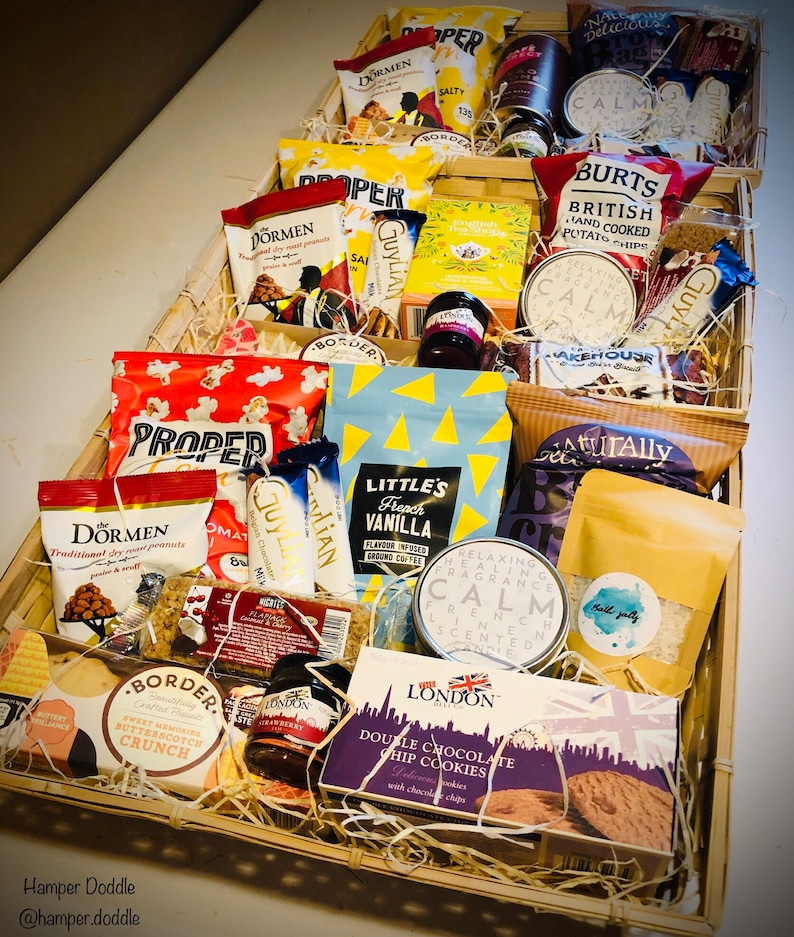 May include: A wicker basket filled with a variety of gourmet food items, including snacks, cookies, and tea. The basket is filled with brown paper shredding and includes brands like Proper, Dormen, and Little's.