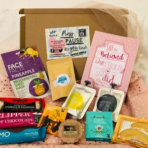 May include: A gift box filled with self-care items. Includes a pineapple face mask, tea bags, a chocolate bar, a card that says "She Believed she Could so she did", and other treats. The box is brown and the items are arranged on a pink polka dot background.