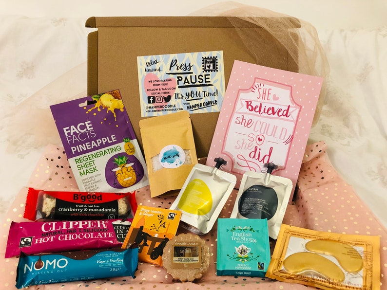 May include: A gift box filled with self-care items. Includes a pineapple face mask, tea bags, a chocolate bar, a card that says "She Believed she Could so she did", and other treats. The box is brown and the items are arranged on a pink polka dot background.