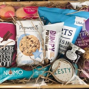 May include: A gift basket filled with various snacks and treats, including a bag of popcorn, a bag of chips, a jar of jam, a box of cookies, a chocolate bar, and a box of instant hot chocolate. The basket is lined with brown paper shreds and has a clear plastic wrap over the top.